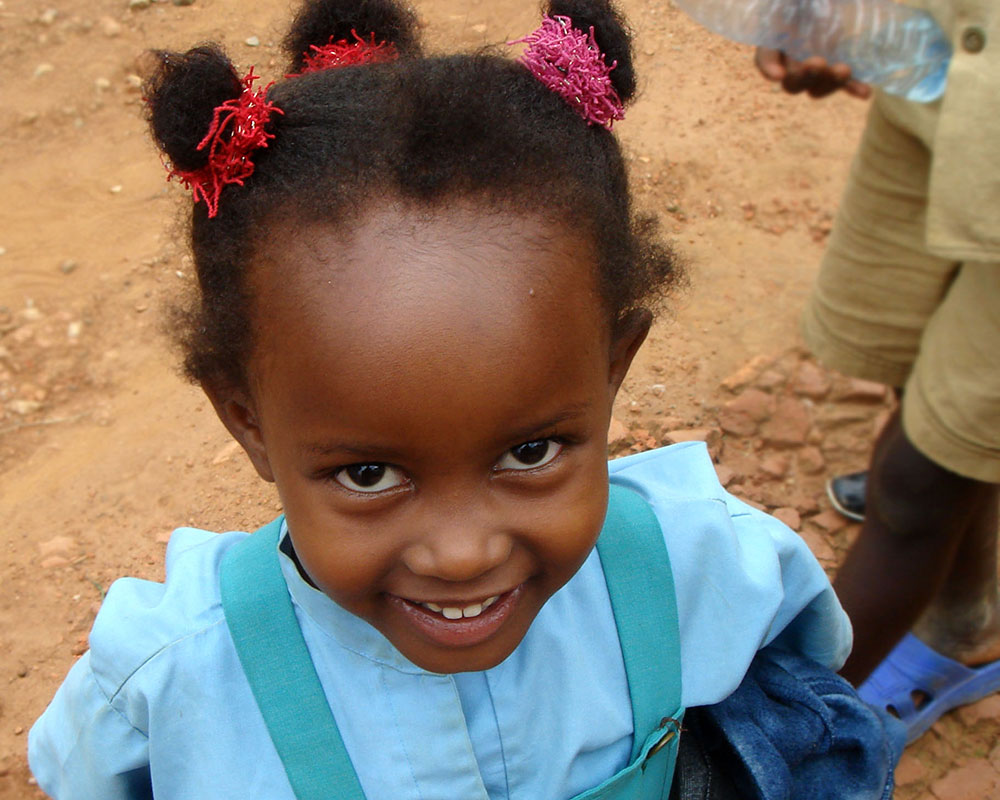 Young Black girl in blue shirt with three little pony tails looking straight up at the camera, smiling.